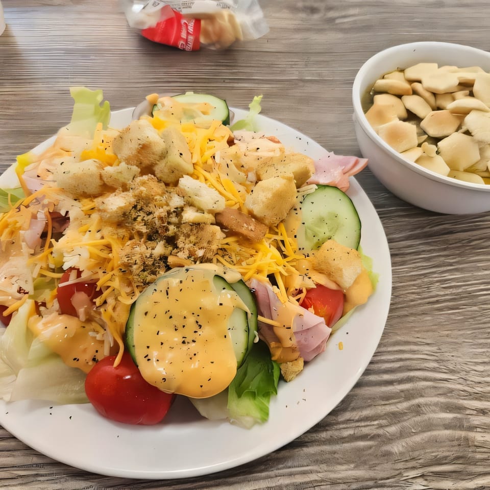 A fresh salad with lettuce, tomatoes, cucumbers, ham, cheese, croutons, and dressing, with a side of oyster crackers.